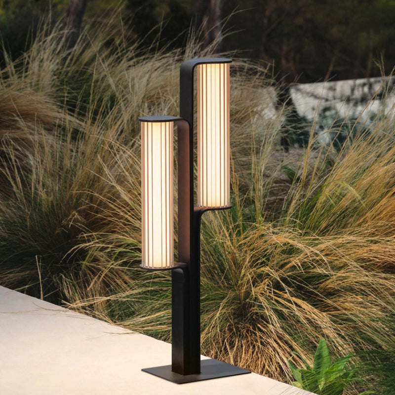 A low-profile outdoor garden lamp with dual striped acrylic shades softly lights up the side of a pathway, surrounded by wild grass.