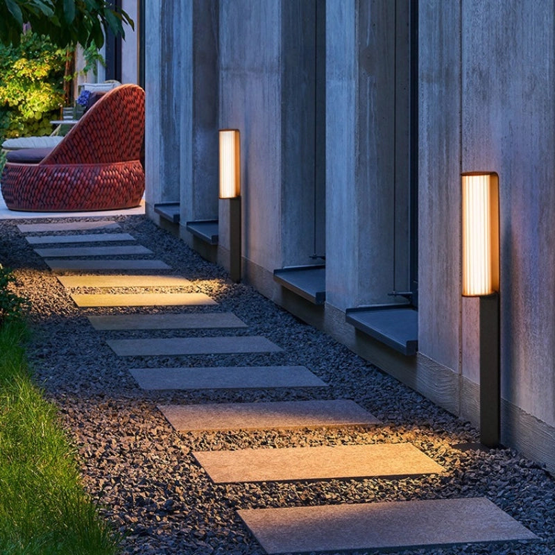A slender ground lamp stands quietly in the outdoor landscape, its warm yellow light spilling gently across a gravel path, creating a calm and inviting nighttime ambience.