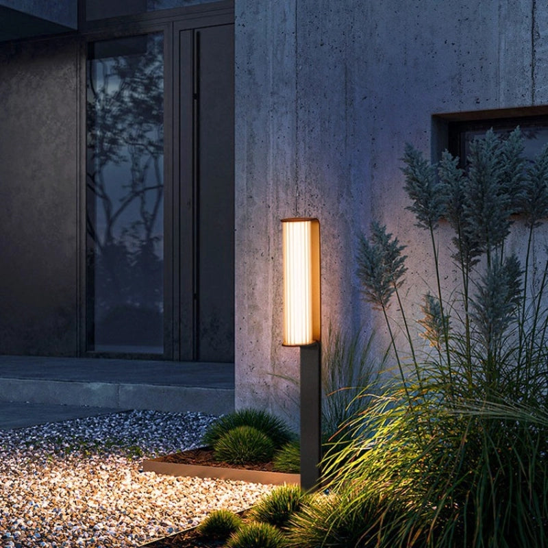 The lamp features a slim, rectangular column design with a dark-toned body. Its warm yellow light softly radiates from the illuminated surface, gently brightening the surroundings. Paired with modern architectural facades, gravel flooring, and lush greenery, it creates a serene and elegant nighttime atmosphere—blending functional lighting with decorative appeal.