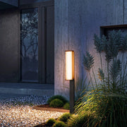 The lamp features a slim, rectangular column design with a dark-toned body. Its warm yellow light softly radiates from the illuminated surface, gently brightening the surroundings. Paired with modern architectural facades, gravel flooring, and lush greenery, it creates a serene and elegant nighttime atmosphere—blending functional lighting with decorative appeal.