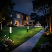 A modern-style villa is located in the center, with lights coming from the interior. There are several columnar floor lamps standing on the lawn, illuminating the lawn with warm yellow light. The path is winding and dotted with flowers and plants. There is a fountain in the distance.