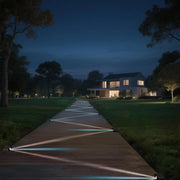 The outdoor scene at night, a wooden path is illuminated by lights on both sides, and the lights project geometric patterns on the ground, leading to a modern-style house surrounded by green trees and grass, creating a peaceful and warm atmosphere.
